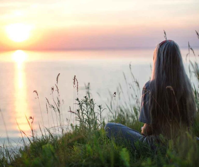 Vrouw zit aan het water en kijkt uit over het landschap bij zonsondergang.
